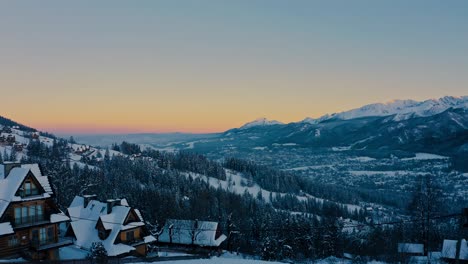 Stunning-Drone-Aerial-Shot-during-sunset-above-winter-trees-and-houses-covered-in-snow
