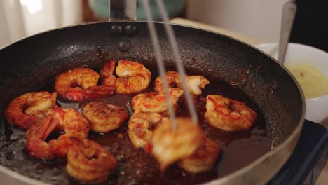 Cooked-prawns-added-to-a-sizzling-pan-with-a-red-seasoning,-lifted-with-metal-chopsticks-to-swirl-and-prevent-burning