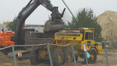 Intensive-work-on-the-construction-site-located-near-the-coast-of-the-sea,-loader-fills-the-truck,-white-sand-piles,-overcast-day-with-fog,-distant-medium-shot