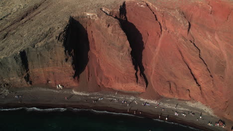 Luftaufnahme-Des-Roten-Strandes,-Insel-Santorini,-Griechenland,-Menschen-An-Der-Sandküste-Unter-Steilen-Klippen