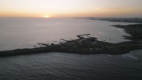 Aerial-Drone-Fly-Above-Peninsula-of-Punta-Carretas-Uruguay-at-Sunset,-Golden-Skyline-and-Calming-Atlantic-Ocean-Waves