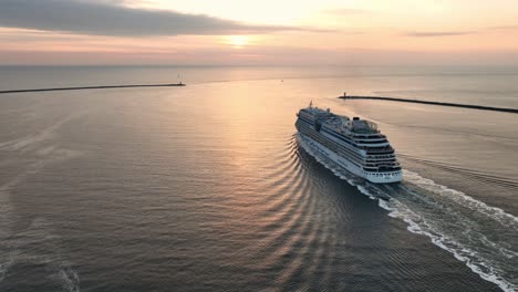 Stunning-aerial-panning-shot-of-a-cruise-shipping-steaming-out-of-a-harbor-into-open-water-with-the-sun-setting-on-the-horizon