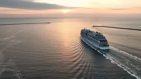 Atemberaubende-Schwenkaufnahme-Aus-Der-Luft-Eines-Kreuzfahrtschiffes,-Das-Aus-Einem-Hafen-Ins-Offene-Wasser-Dampft,-Während-Die-Sonne-Am-Horizont-Untergeht