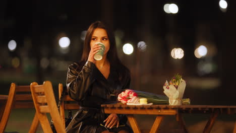 Pretty-Brunette-Elegant-Woman-Sitting-Alone-At-Cafe-Terrace-By-Night-Drinking-Coffee,-Cinematic-Bokeh