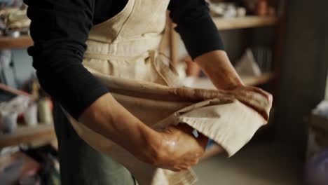 Young-happy-potter-wipes-his-hands-on-his-apron-after-working-with-clay.-Close-up.-The-concept-of-traditional-professions
