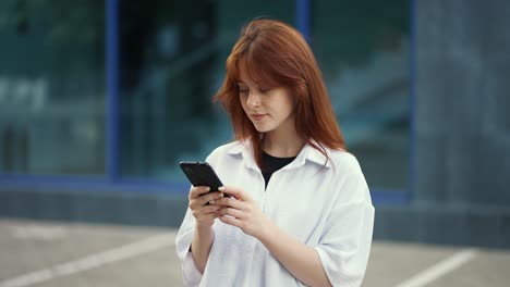 a-red-haired-girl-in-a-shirt-is-typing-text-on-the-phone-against-the-background-of-a-city-building