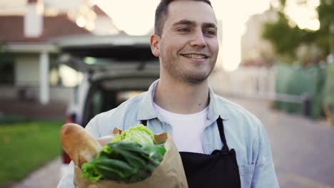 Close-up-handsome-smiling-courier-man-going-from-the-car-to-house-and-holding-paper-bag-with-food-outdoors.-Worker-grocery