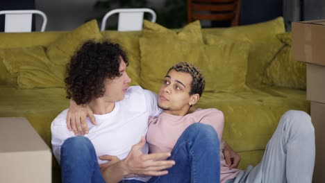 Homosexual-couple-hugging,-talking-in-a-new-apartment-around-boxes