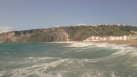 Low-aerial-shot-overhead-waves-crashing-onto-Nazare-beach-surrounded-by-cliffs