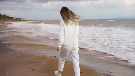 Blonde-woman-walking-along-ocean-coast-alone,-rear-view