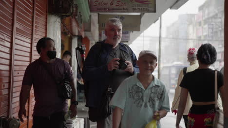Old-Male-Photographer-Taking-Photos-On-The-Sidewalk-With-Crowd-Of-Passersby-On-A-Rainy-Day