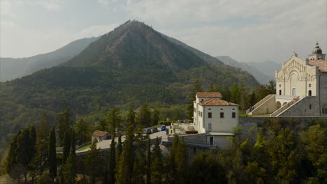 Spectacular-aerial-dolly-of-mountaintop-village-and-church,-Sanctuary-of-Montecastello,-with-a-mountainous-and-forest-covered-background