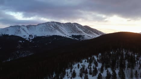 Aerial-view-of-a-dramatic,-cloudy-sunset-behind-a-snowy-mountain-range,-in-Colorado