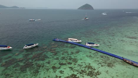 Clear-turquoise-water-of-the-Andaman-Sea,-coral-and-underwater-structure-near-shore,-floating-dock-and-snorkel-trip-boats
