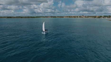 Exciting-Drone-Shot-of-a-Sailing-Yacht-Leaving-the-Shoreline-and-Venturing-into-the-Vast-Ocean,-Revealing-a-Spectacular-View-of-the-Surrounding-Beach,-Green-Jungle-and-Picturesque-Holiday-Apartments
