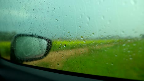 Ein-Heftiger-Regenschauer,-Der-Aus-Dem-Autoinneren-Betrachtet-Wird,-Mit-Regentropfen,-Die-über-Die-Windschutzscheibe-Fallen,-Und-üppigen-Grünen-Feldern-Dahinter