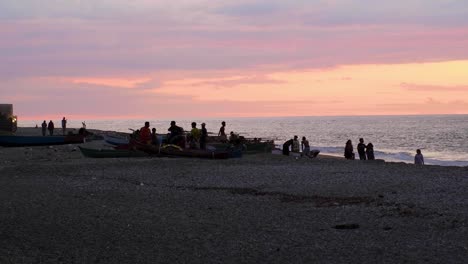 People-relaxing-and-socialising-near-fishing-boats-on-Beto-Tasi-beach-during-pink-sunset-in-capital-Dili,-Timor-Leste,-Southeast-Asia