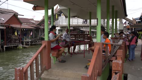 Locals-sit-under-the-pavilion-with-the-pythons-while-waiting-to-offer-tourists-a-memorable-photo-opportunity-in-Damnoen-Saduak-Floating-Market,-Ratchaburi-province,-South-West-of-Bangkok,-Thailand