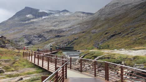 Tourists-walking-on-a-fenced-walkway-across-uneven-terrain-next-to-a-mountain-river,-Trollstigen-visitors-center-and-Norwegian-mountains-in-background,-handheld-dolly-shot,-tourism-and-travel-concept