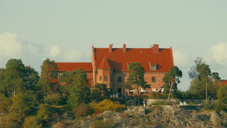 An-aerial-view-of-a-gathering-taking-place-in-the-backyard-of-a-large-red-brick-manor-home-on-a-hill-with-few-trees-growing-nearby,-filmed-in-UHD-4K