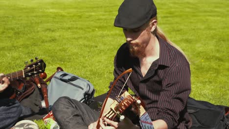 Bearded-man-with-long-hair-plays-a-nyckelharpa,-also-called-a-key-fiddle-or-a-Scandinavian-violin,-outside-sitting-on-the-grass