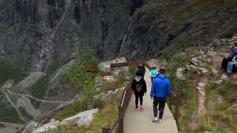 Tourists-walk-down-a-paved-pathway-on-the-edge-of-a-mountain-towards-a-jutting-viewpoint-at-Trollstigen-in-Norway,-overlooking-the-winding-road-and-waterfall-in-the-background