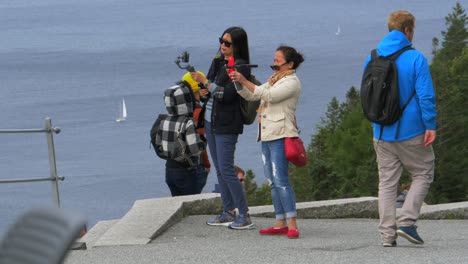 Sailboats-on-the-water-in-the-distance-as-two-Asian-women-take-pictures-using-their-phones-and-smartphone-gimbals