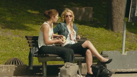 Two-attractive-young-women-sit-on-a-park-bench-talking-and-eating-apples-in-the-sunshine