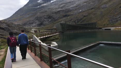Pareja-Amorosa,-Tomados-De-La-Mano-Y-Caminando-Por-Un-Camino-Concreto-En-Trollstigen-En-Noruega,-El-Centro-De-Visitantes-Y-Las-Altas-Montañas-En-El-Fondo,-Concepto-De-Viajes-Y-Turismo,-Foto-De-Seguimiento-Manual