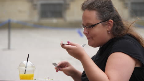 Obese-woman-munching-on-desserts-with-juice-in-a-outdoor-cafe,-slow-motion-shot