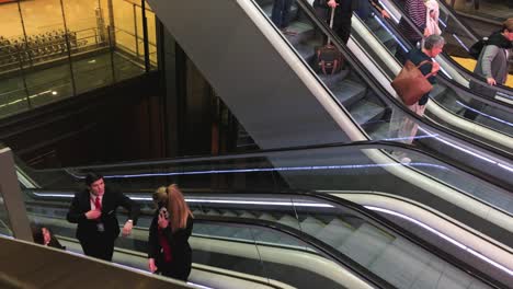 People-stepping-off-a-two-row,-parallel-escalator-staircase,-towing-carry-on-luggage,-busy-airport-terminal-scene,-passengers-rushing,-travel,-business-and-transportation-concept