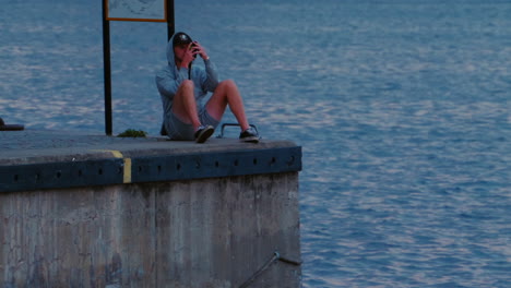 Young-caucasian-man-looking-at-phone-sitting-on-pier,-evening