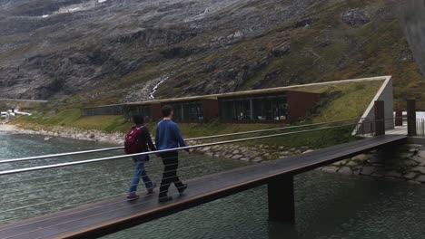 Mid-aged-couple,-holding-hands-and-crossing-a-bridge-across-a-mountain-river-on-Trollstigen-pathway-in-Norway,-the-visitors-center-and-high-mountains-in-background,-handheld-shot,-love-and-couples