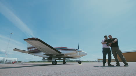 Two-men-standing-on-the-tarmac-to-the-right-of-a-small-airplane-discussing-while-checking-a-computer