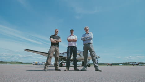 Three-men-standing-on-the-tarmac-in-front-of-a-small-airplane-with-other-airplanes-and-the-blue-sky-in-the-background