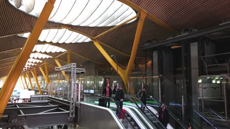 Pasajeros-Con-Equipaje-De-Mano-Subiendo-Escaleras-Mecánicas-En-Un-Edificio-Moderno,-Terminal-Principal-Del-Aeropuerto-Internacional-De-Madrid-Barajas-Con-Su-Moderno-Diseño-Arquitectónico,-Concepto-De-Viaje-Aéreo