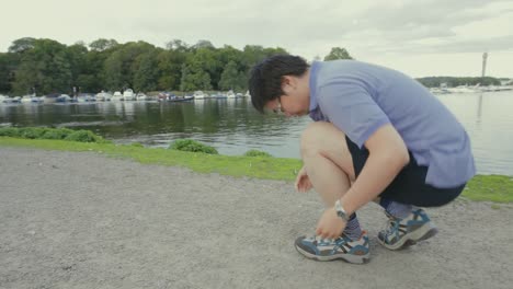 Young-Asian-man-in-a-purple-shirt-tying-his-shoe-near-a-lake-then-begins-to-run
