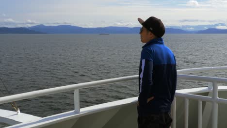 Asian-man-with-baseball-cap-and-jacket-standing-outside-near-the-bow-of-a-ferry-boat-transiting-a-Norwegian-fjord-looks-out-at-the-landscape-on-the-horizon