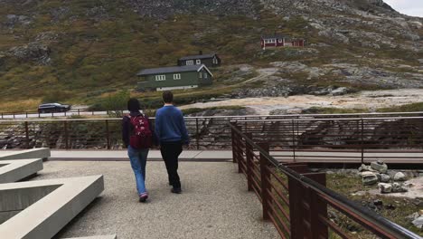 A-couple,-holding-hands-and-walking-down-a-concrete-pathway-at-Trollstigen-in-Norway,-the-visitors-center-and-high-mountains-in-background,-travel-and-tourism-concept,-handheld-follow-shot
