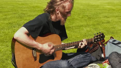 Ein-Junger-Mann-In-Jeans-Und-Schwarzem-Hemd-Spielt-Akustikgitarre,-Während-Er-Draußen-Im-Gras-Sitzt