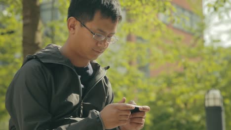 A-young-Asian-man-wearing-a-jacket-sitting-outside-playing-a-game-on-his-phone