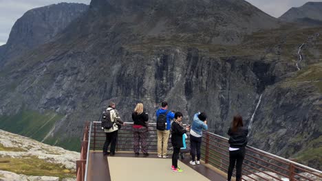 Tourists-on-a-modern-viewpoint-in-the-mountains-taking-pictures-of-waterfall-and-mountains-in-front-of-them,-modern-viewpoint-jutting-over-the-edge-of-cliffs-at-Trollstigen-in-Norway