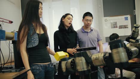 Asiatische-Studenten-Sprechen-Mit-Ihrem-Professor-In-Einem-Robotiklabor,-Während-Sie-über-Ein-Gerät-Diskutieren