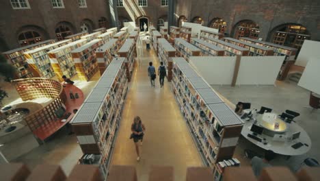 Blick-Von-Oben-Auf-Den-Boden-Der-Universitätsbibliothek-Und-Die-Symmetrischen-Reihen-Von-Bücherregalen,-Während-Studenten-Lernen-Und-Durch-Die-Gänge-Gehen