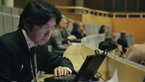 Ein-Asiatischer-Mann-Mit-Brille-Blickt-Während-Einer-Präsentation-In-Einem-Großen,-Modernen-Universitätssaal-Mit-Stadionbestuhlung-Auf-Seinen-Laptop