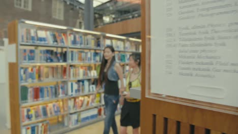 Zwei-Junge-Asiatische-Frauen-Gehen-Durch-Die-Gänge-Der-Universitätsbibliothek-Zwischen-Regalreihen-Voller-Bücher-Zum-Recherchieren-Und-Lernen