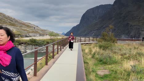 Handheld-dolly-shot-of-tourists-walking-towards-the-camera-at-Troll's-ladder-in-Norway,-an-iron-and-concrete-walkway-next-to-a-mountain-river-with-mountains-in-background,-Trollstigen-in-Norway