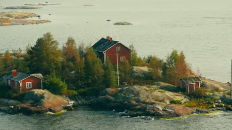 Beautiful-dark-red-wooden-home-and-out-buildings-on-a-small-rocky-island-as-a-sailboat-motors-out-toward-open-water-behind-the-house