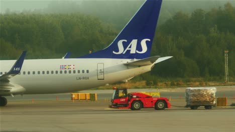 Carro-De-Carga-De-Equipaje-Moviéndose-En-La-Pista-Del-Aeropuerto-De-Estocolmo-En-Suecia,-Junto-A-Un-Avión-De-Pasajeros