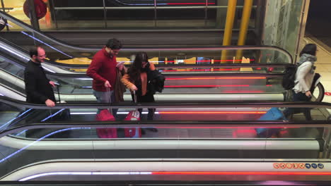 People-stepping-off-a-two-row,-parallel-escalator-staircase,-towing-carry-on-luggage,-busy-airport-terminal-scene,-passengers-rushing,-travel,-business-and-transportation-concept